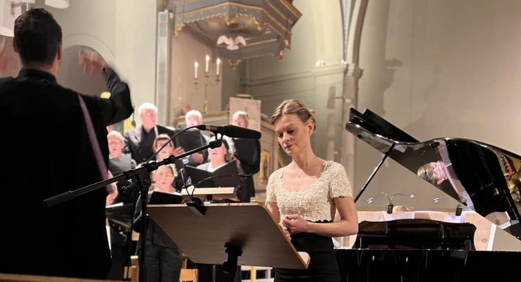 Petra Valman är sopransolist på kyrkokonserten. Står framför flygel med kör och dirigent till vänster.